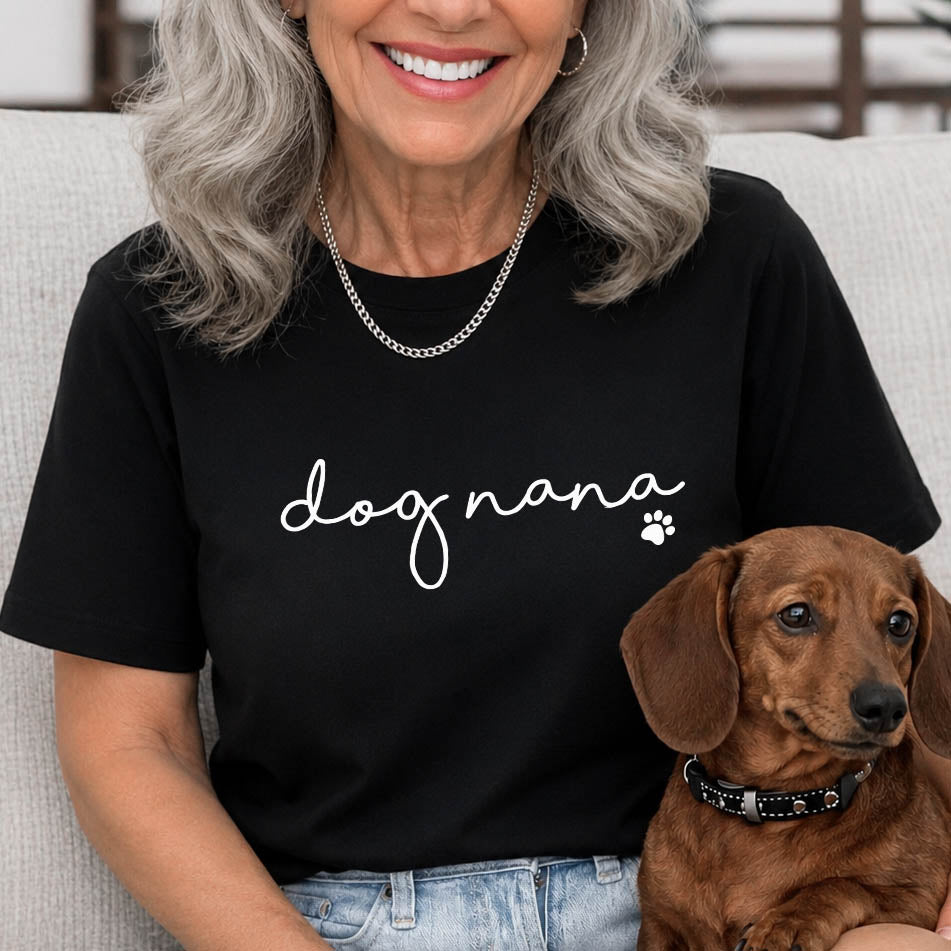 A woman with gray hair wearing a black t-shirt with 'dog nana' written in white script, sitting on a light gray couch with a brown dachshund dog.